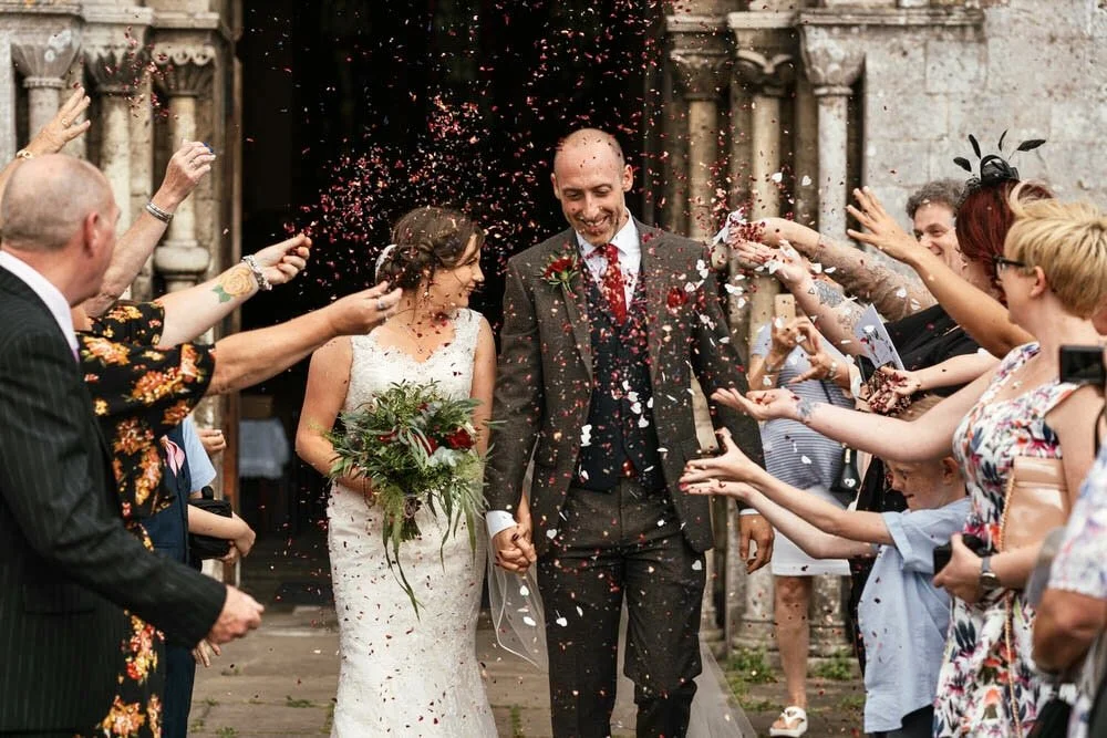 leaving Margam Abbey to a flutter of confetti being thrown from both sides, a happy smiling bride and groom walk down the middle of all their wedding guests. THe groom in a brown tweed suit next to his bride holding a foliage bouquet with red flowers