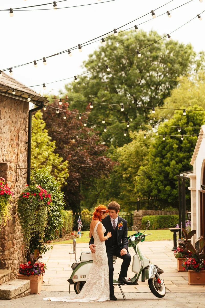 bride and groom posing for their wedding photographs, the groom with a paisley tie and colourful buttonhole is sat on his vespa moped and his wife is leant against him in a lace dress. Surrounded by festoon lighting and trees at the King Arthur hotel