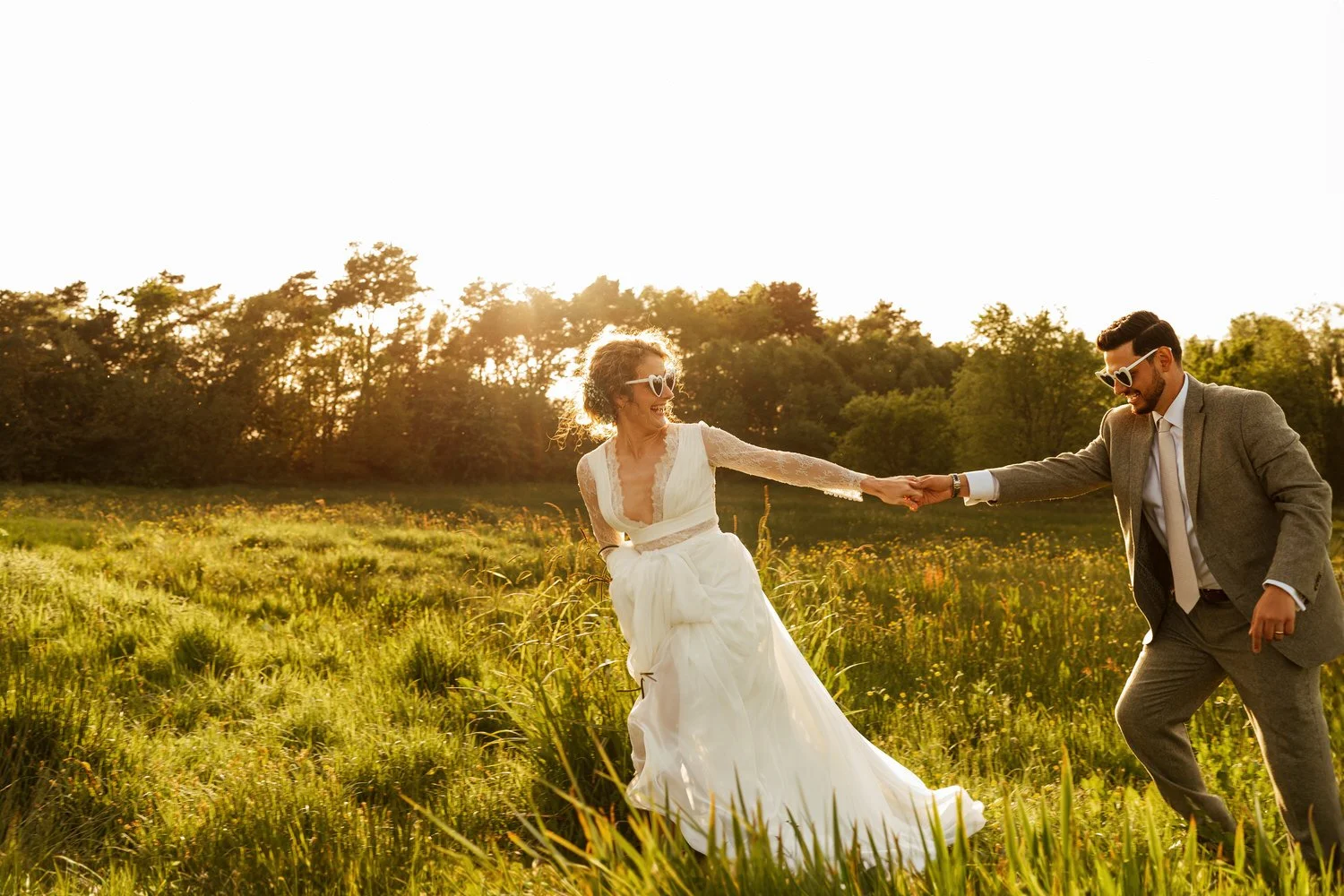 Wedding couple in white heart sunglasses running through a field lit by a setting sun in Swansea.