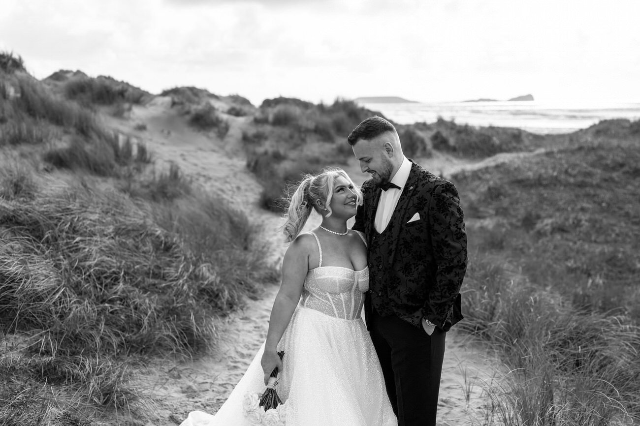 Glamourous bride looking up at her new husband in a black tuxedo on the sand dunes at Llangennith, with Worm's head Rhossili in the background.