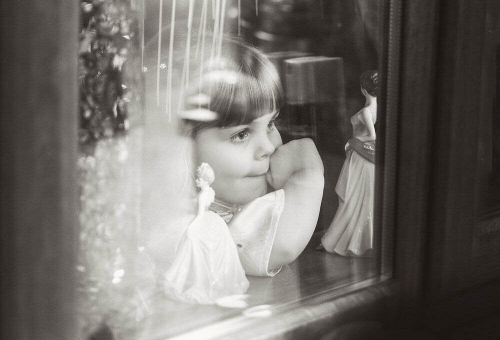 Young flower girl looking out of a window on the wedding day. Surrounded by figurines and a very cute face being pulled.