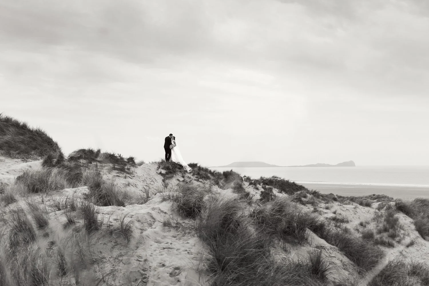 Black and white image of a wedding couple kissing on the sanddunes at Llangennith, Worms head and Rhossili beach in the background of the newlywed couple during their portraits for their wedding film.