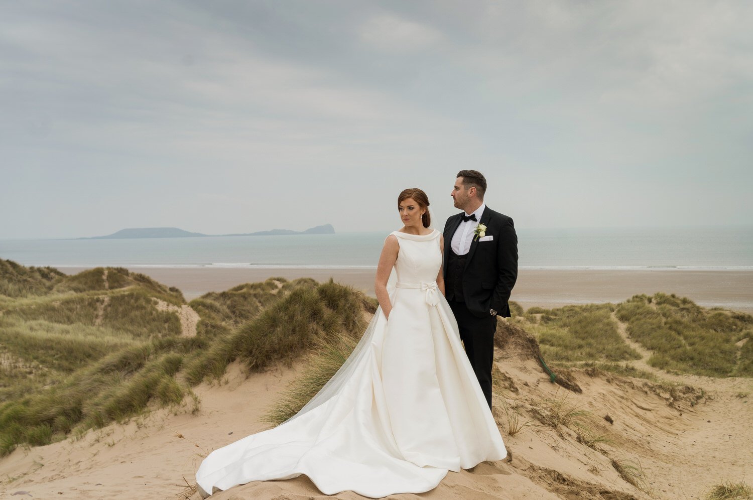 Sophisticated wedding photography at Llangennith sand dunes with Worms head in the background. A newly married wedding couple in the foreground, the bride with her hands in the pockets of her wedding dress and husband in a black tuxedo.