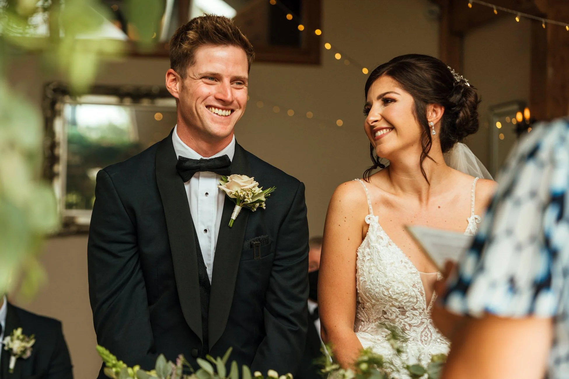 A wedding photograph taken at Oldwalls venue in Swansea featuring a groom on the left in a black and navy tuxedo with black bow tie, he is smiling and his bride on his right in an ivory lace, she has dark hair in an up do. Fairy lights behind.