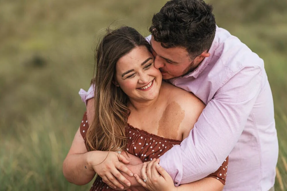 An engaged couple embrace and laugh naturally during a pre wedding photo shoot at Pembrey, Surrounded by long grass in the background.