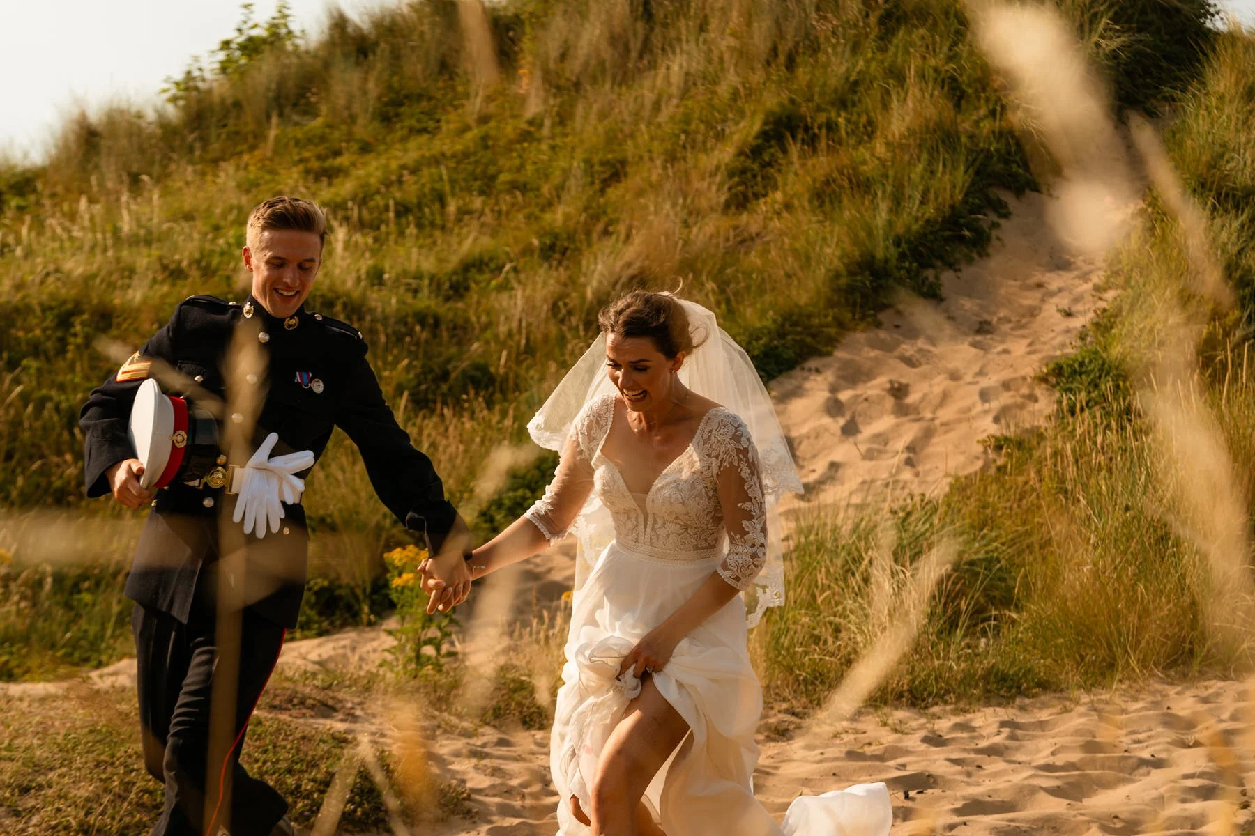 Newly married couple running down a sand-dune at Oxwich bay after their ceremony. The groom is laughing, holding his Royal Marines hat, wearing his number 1s military uniform, the bride in a lace detailed dress.