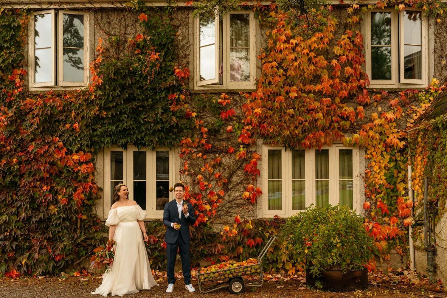 Swansea wedding at Parc Le Breos, a bride holding an autumn colour bouquet of flowers and wearing a gold crown, leaning as she laughs looking at her groom next to her juggling apples. Autumnal leaf covered building with an apple cart in front.
