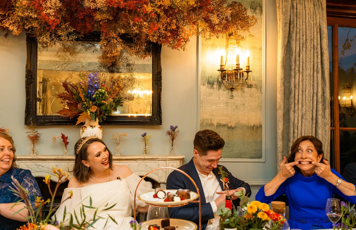 Funny moment during wedding speech as the mother of the groom in a royal blue suit reenacts a memory of her son the groom, sat with the bride & groom at the top table with a floral display of autumn decor behind in a Parc Le Breos wedding in Swansea