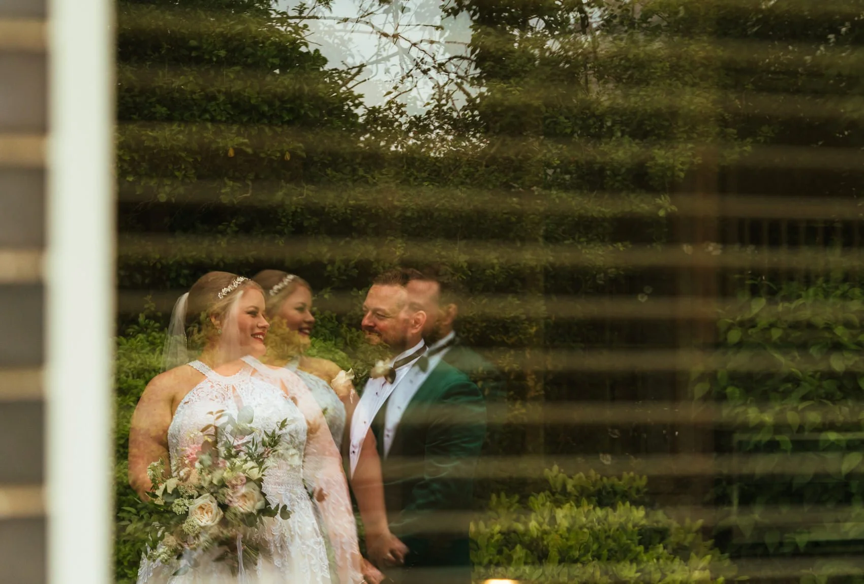 Window reflection of a bride and groom looking towards each other smiling, bride in a halterneck dress holding a bouquet with her groom in an emerald green tuxedo jacket surrounded by abstract greenery and reflections.