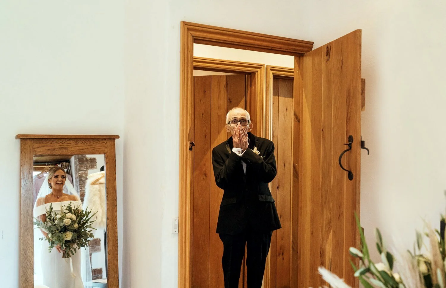 Father of bride with hands over his face in shock reaction to seeing his daughter in her wedding dress for the first time at Rosedew farm in Llantwit Major. Father in door way, bride in mirror reflection smiling holding her ivory and green bouquet.