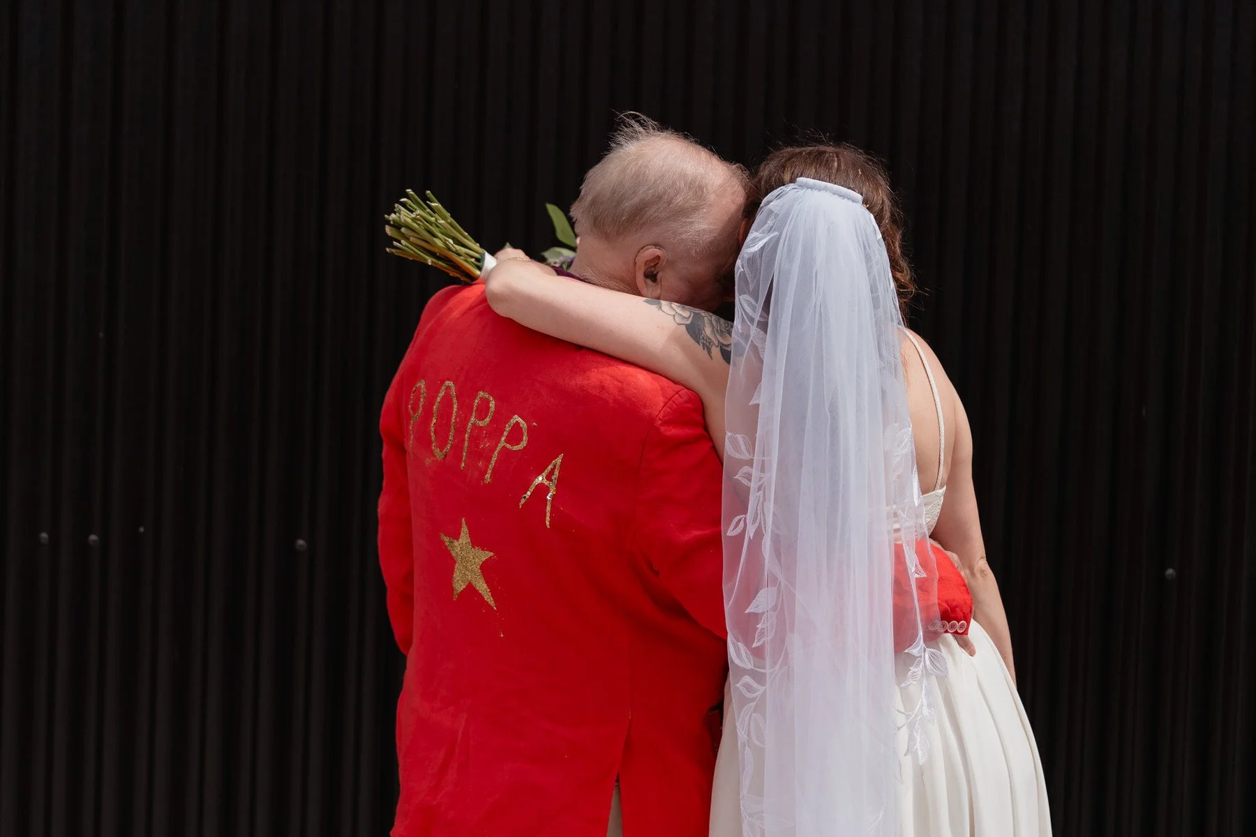 Bride in white dress hugs man in red jacket labeled POPPA with a yellow star, embracing after the shack revolution wedding ceremony; both face away.