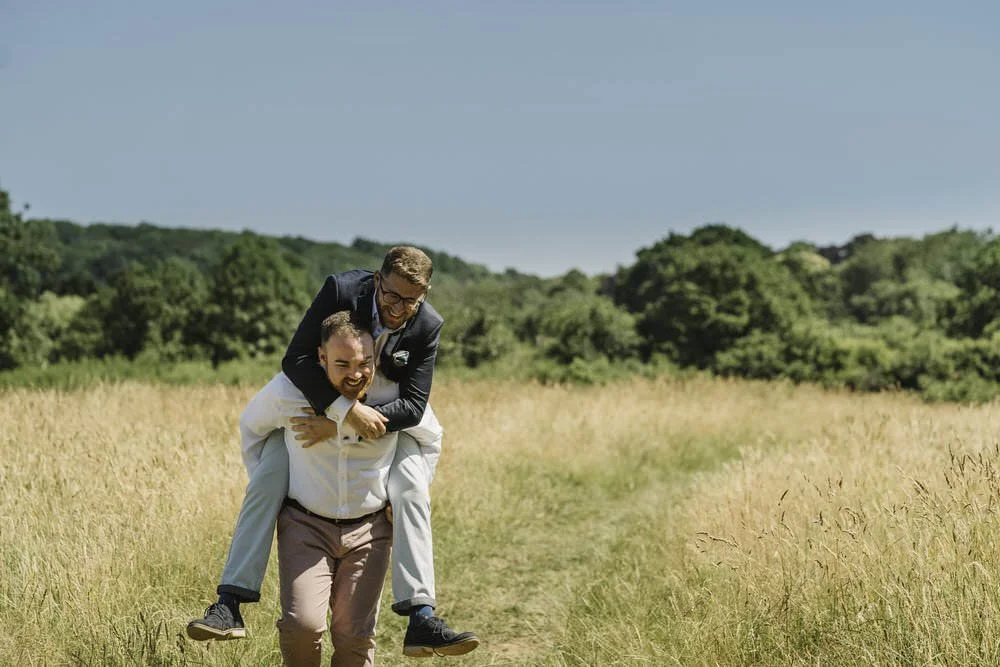 Happy engagement photo of two men running through a nature reserve in St Fagans, having a fun piggy back for their engagement photo shoot.