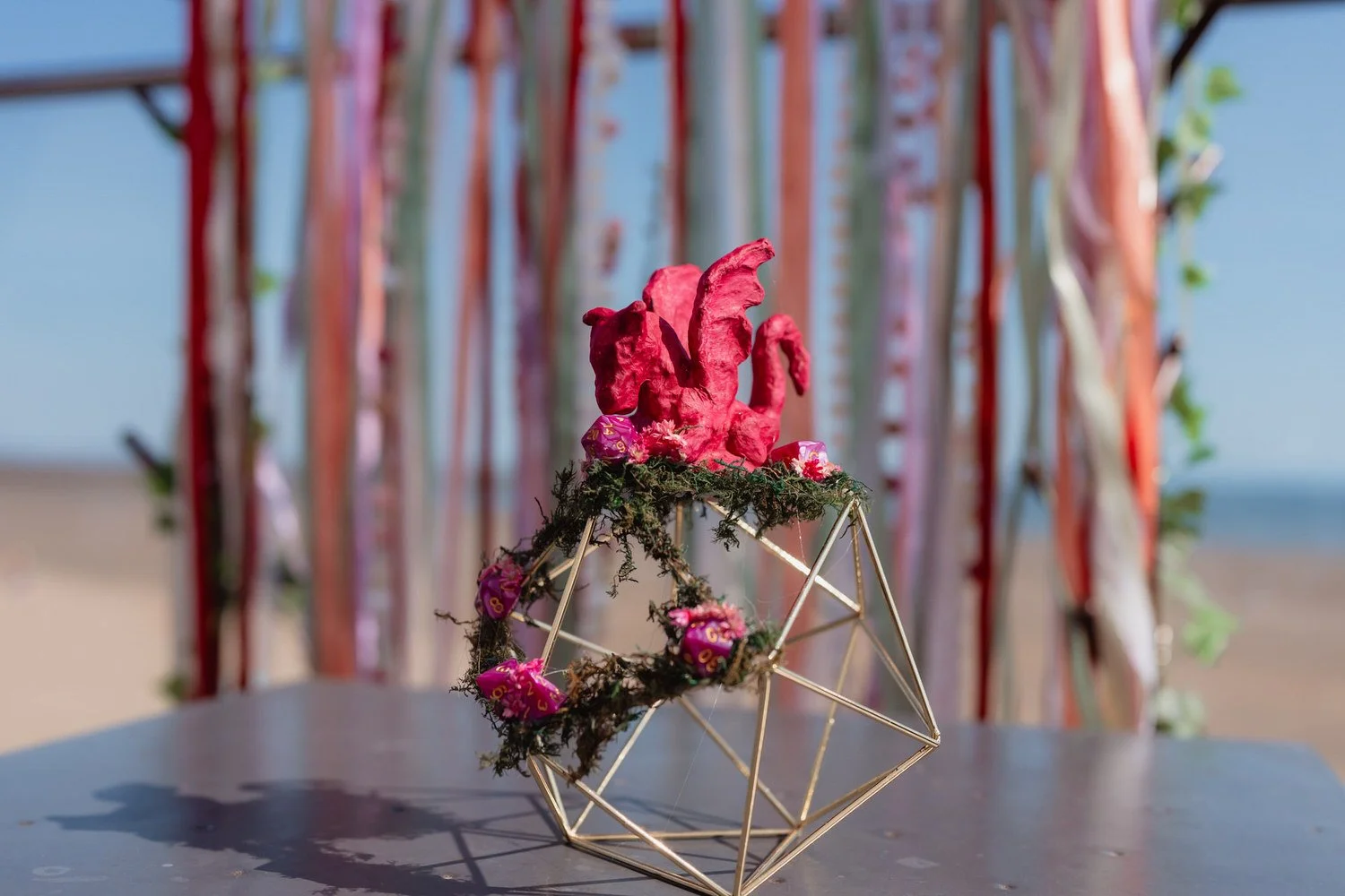 Wedding table decoration, a Dungeons and Dragons themed structure made of a golden shape with a red dragon on top. Background of hanging multi coloured ribbons