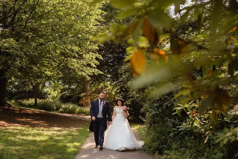 Newly married wedding couple in wedding attire walking through Clyne Gardens in Swansea. Surrounded by greenery and trees.