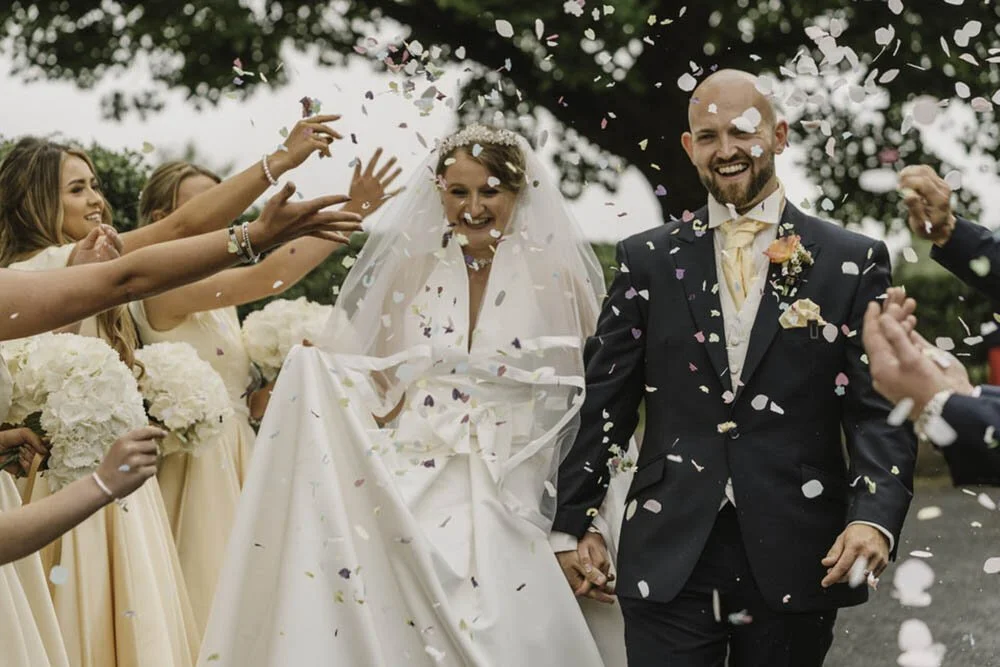 Young happy couple just married, walking though fluttering confetti. Wedding guests throwing confetti over the newlyweds outside Samlet Church in Swansea.