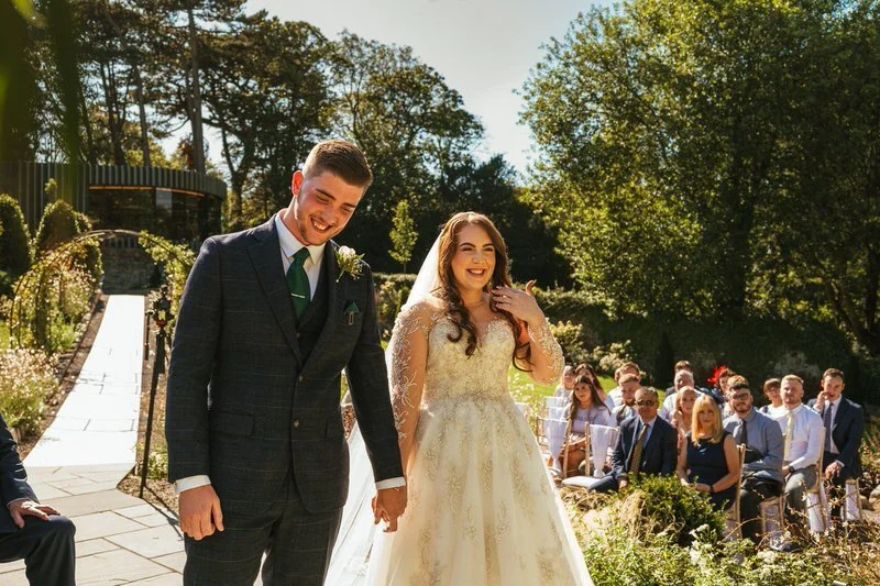 young bride and groom nervously laughing during their outdoor ceremony at Fairyhill wedding venue, surrounding by forestry and sunshine the groom has a green tie and pocket sqaure. the bride has a detailed lace gown and veil.