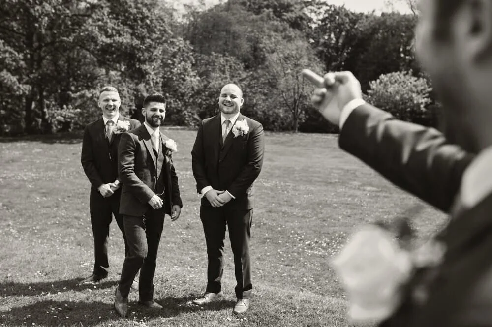 Smart looking groom and his groomsmen, shown in black and white, laughing and joking in a relaxed natural moment, shot past the arm of another groomsmen who they are laughing at because he is flicking the bird, taken at Bryngarw House.