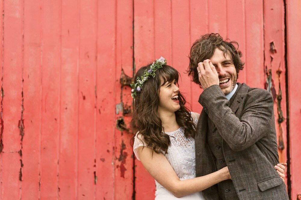 Indie looking alternative bride and groom laughing naturally in front of a rustic battered old red wooden set of doors. Bride with dark curly hair in a simple lilac and gyp flower crown, at Mumbles Pier.