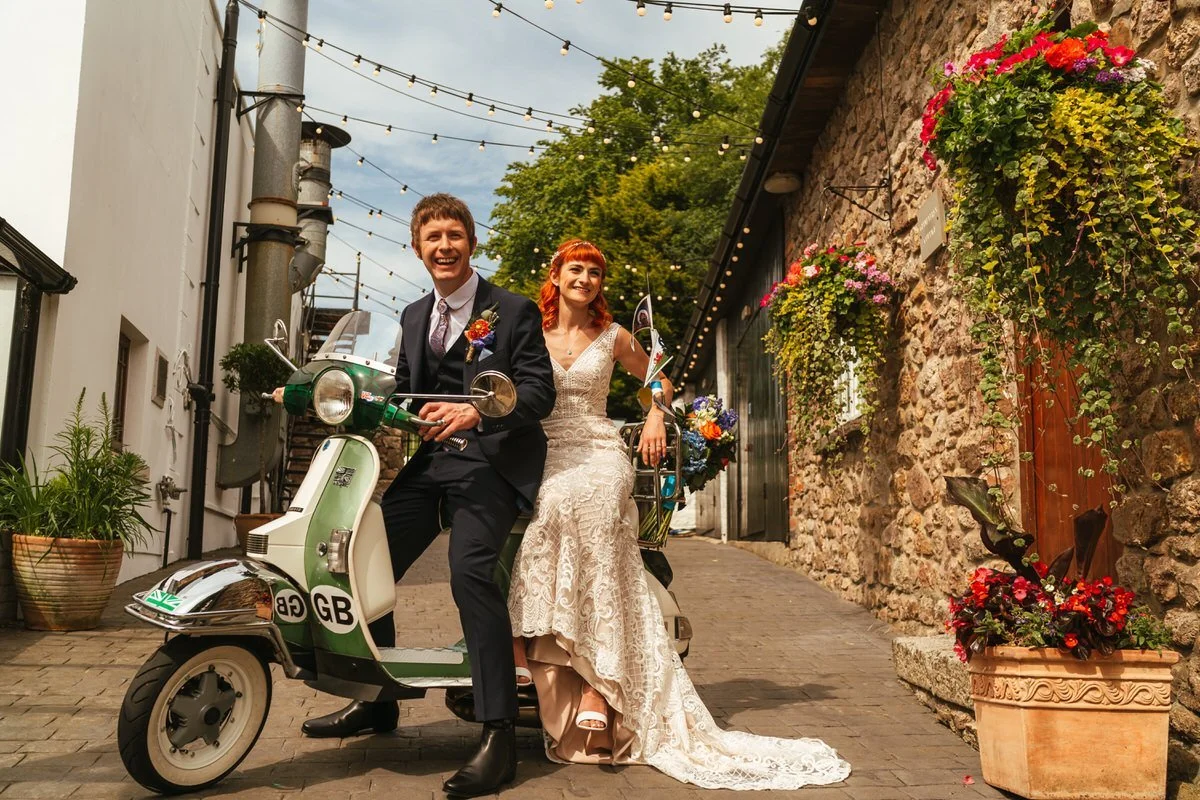 Bride and groom with a mods and rockers themed look to their wedding, sat outside in the courtyard of the King Arthur Hotel, sat on a green vintage vespa laughing naturally into the distance. The bride's bouquet of colourful flowers on the back rack.