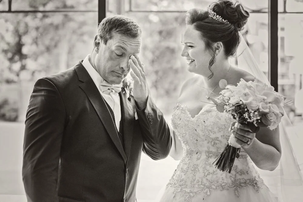 Groom crying at first look of his bride, taken before their wedding ceremony in Swansea. The bride who is laughing, holding her bouquet of flowers as she looks on at her groom rubbing his eyes.