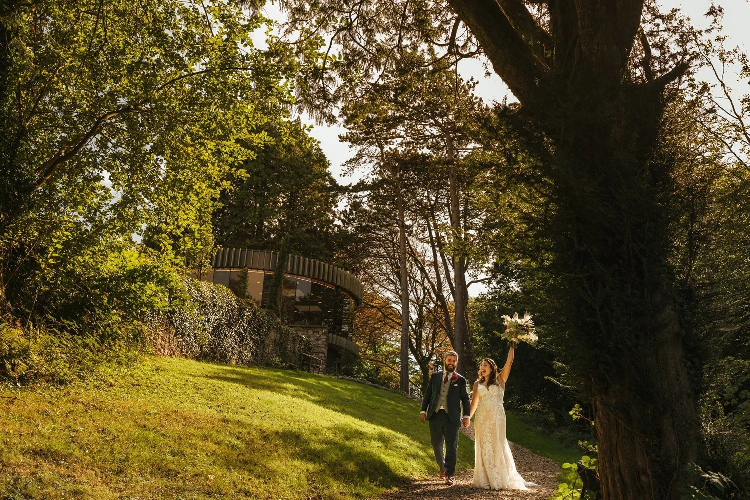 bride and groom walking hand in hand laughing, the bride has her bouquet raised into the air excitedly. Behind them is the glass K room at Fairyhill wedding venue in Swansea. Taken during their wedding couples pictures.