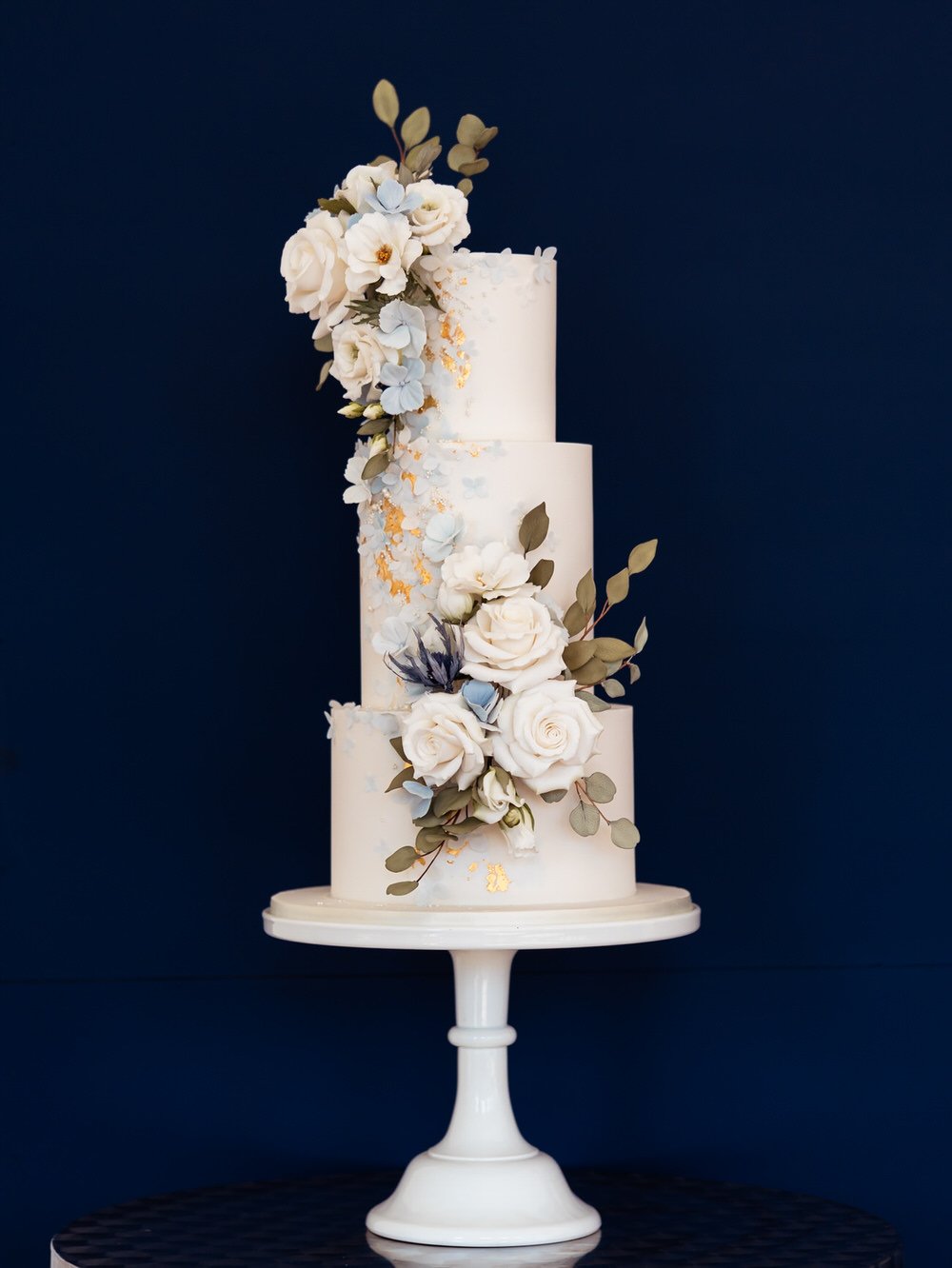 Navy background with a Gold leaf wedding cake decorated in cream flowers, set against a deep navy background.