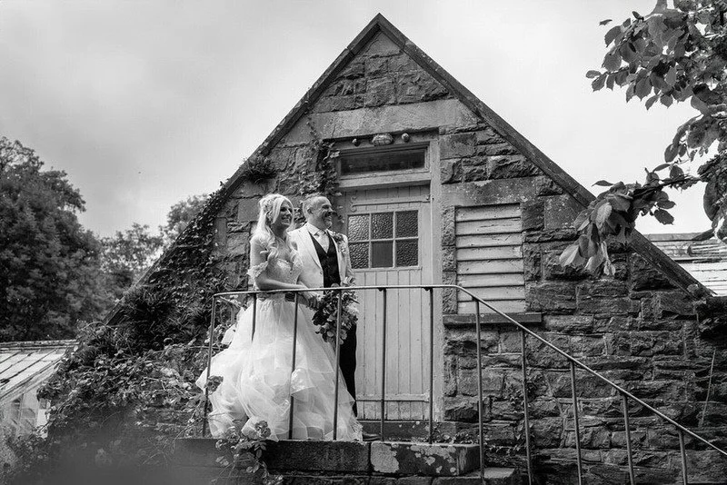Black and white wedding photograph of a happy couple outside an old stone barn, looking outwards smiling in to the distance. Taken at Plas Dinam Country House in Mid Wales.