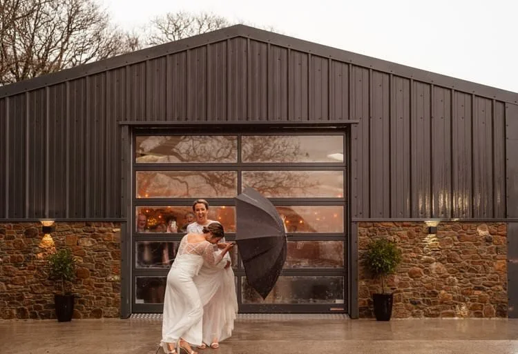 Two brides in dresses laughing and walking under a clear umbrella at West Regwm Farm in West Wales.