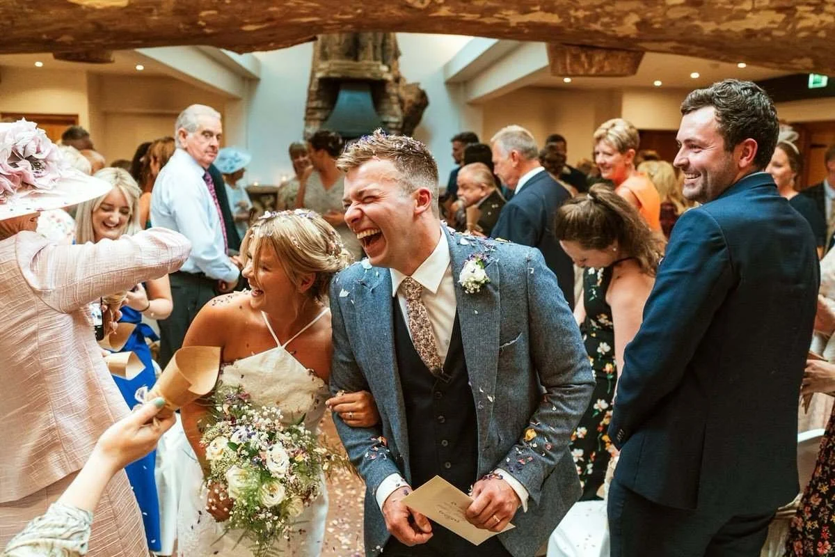 A bride and groom laughing joyfully during their wedding celebration at the Oxwich bay hotel, surrounded by guests in a warmly lit indoor venue. The bride holds a bouquet of flowers, and the groom is holding a card.