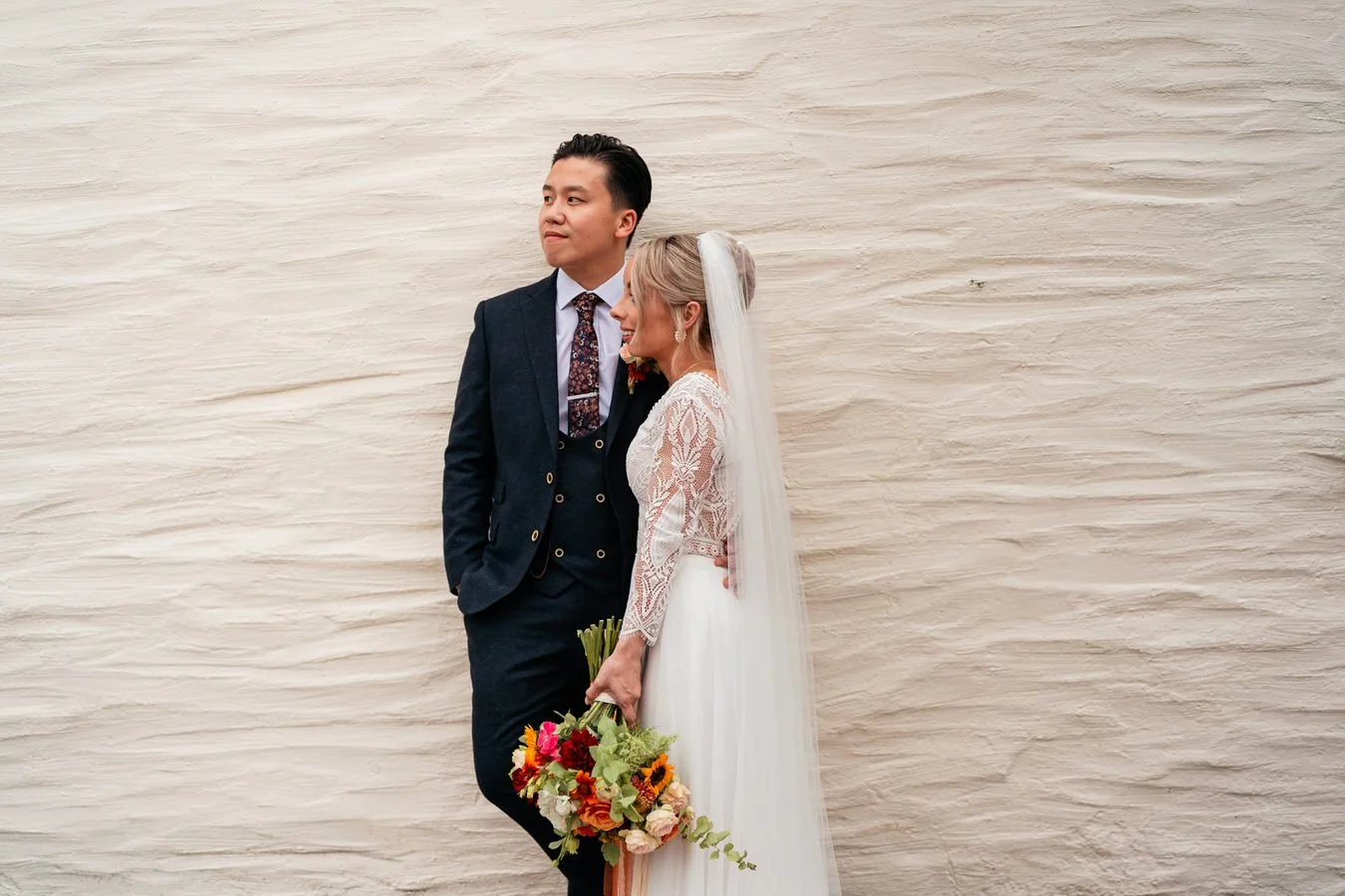 Cool young couple leaning against a wall on their wedding day during their film being created. Bride holding flowers.