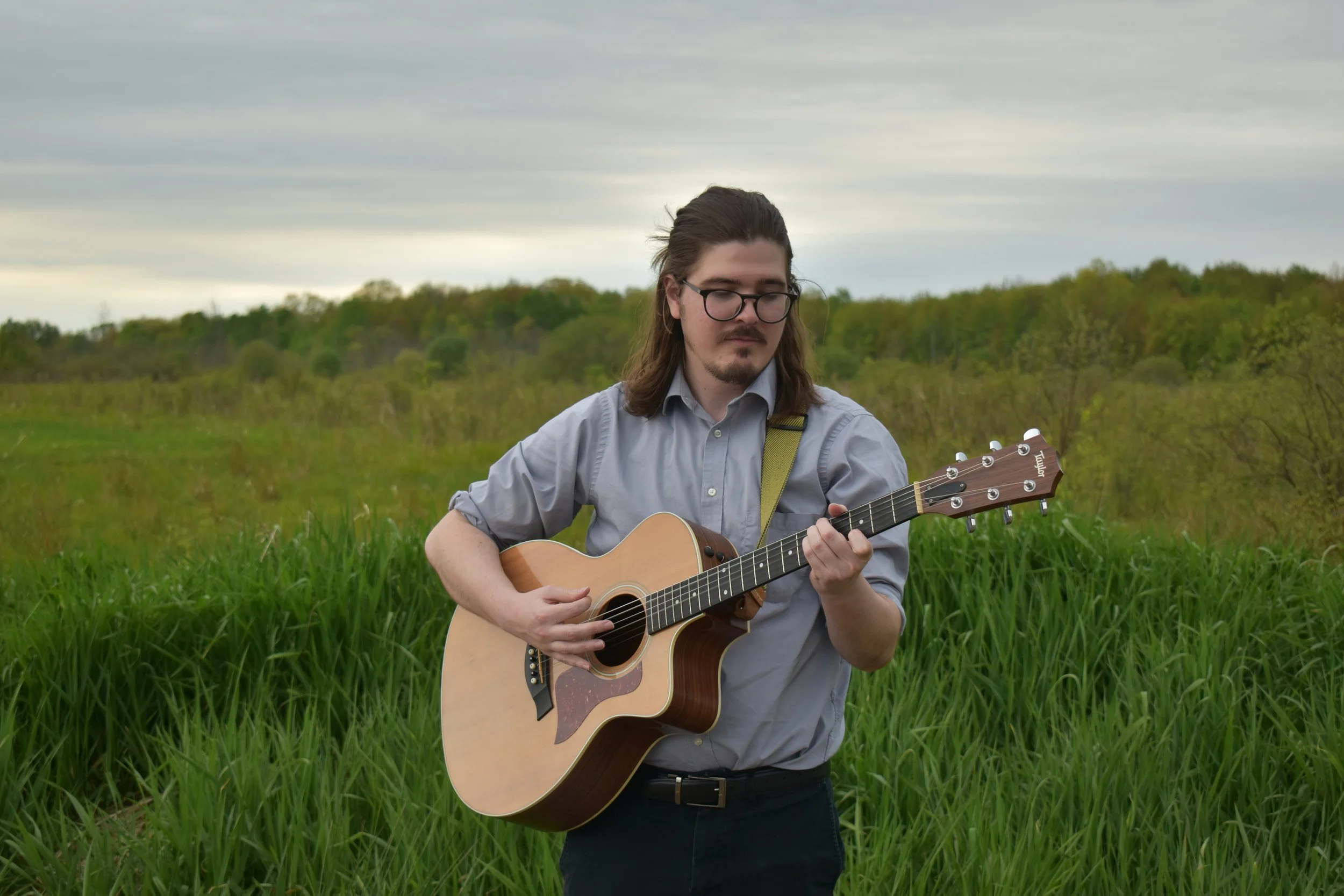 A man with long hair and glasses playing an acoustic guitar outdoors in a grassy field with trees and cloudy sky in the background.