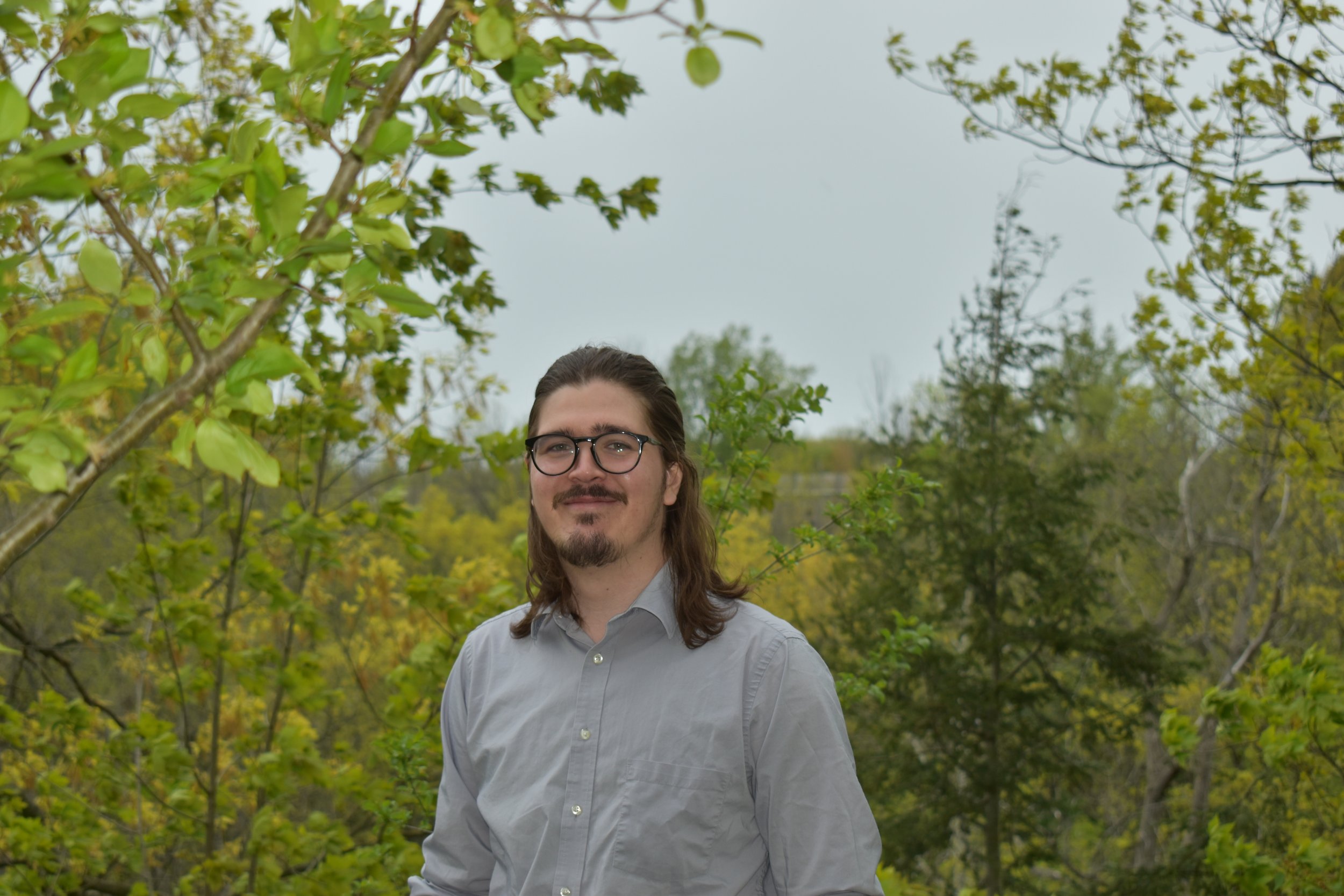 A man with glasses and long hair smiling outdoors among green trees.