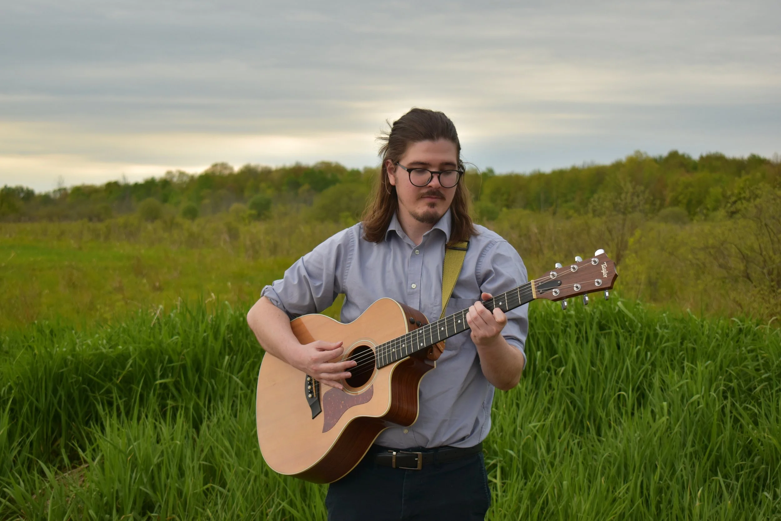 A young man with long hair and glasses playing an acoustic guitar in a grassy field with green trees and an overcast sky in the background.