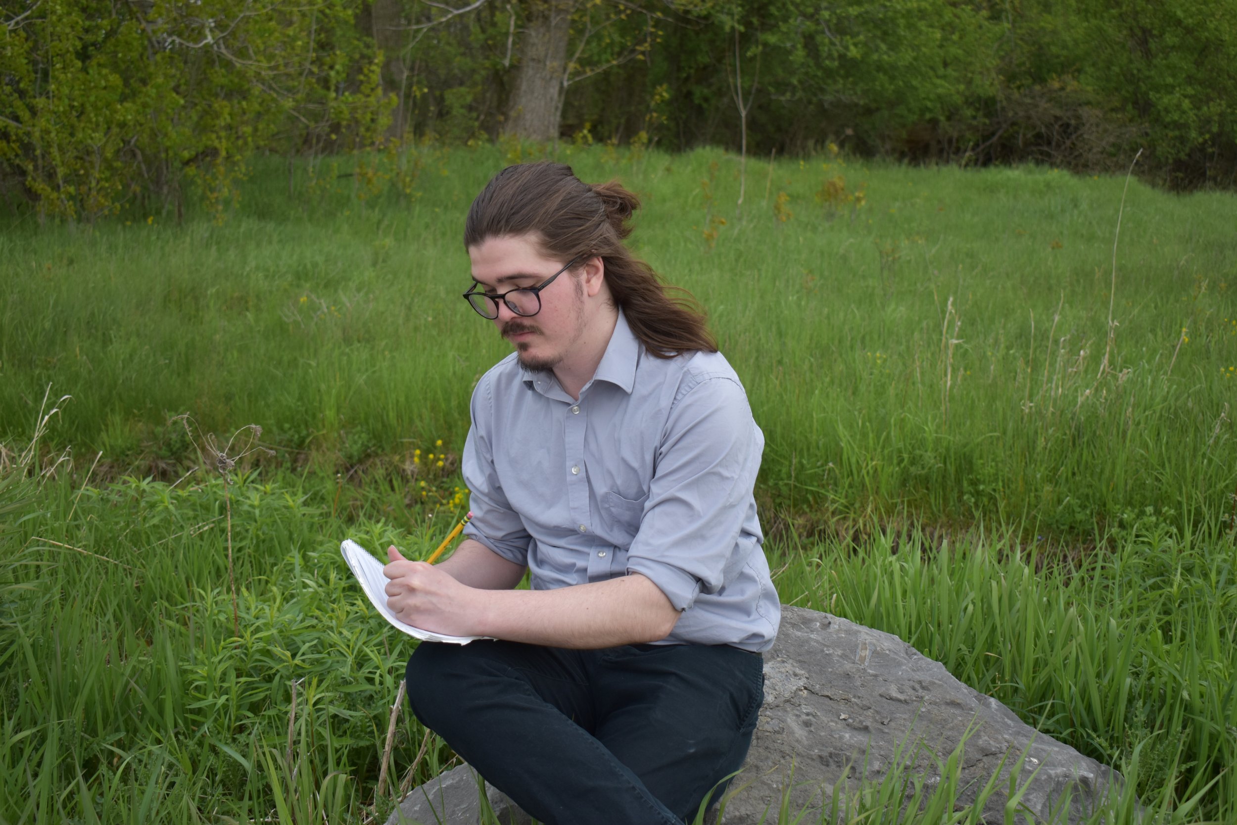 A man with long hair, glasses, and a light gray shirt sitting on a rock in a grassy outdoor area, writing in a notebook with a yellow pencil.