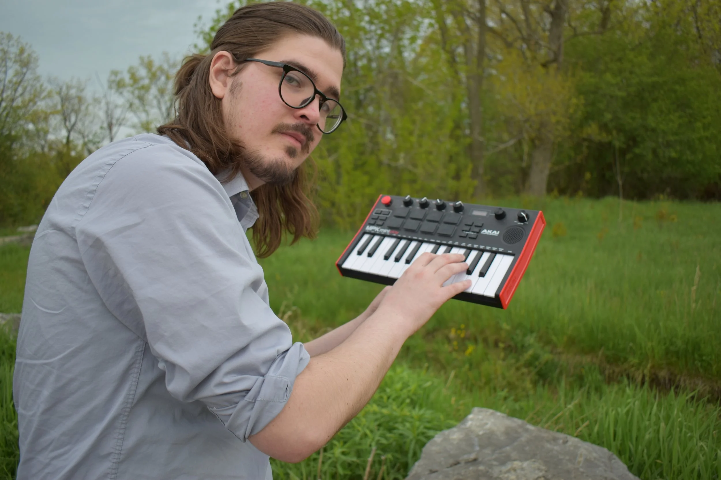 A young man with long hair and glasses playing a small keyboard outdoors in a green park.