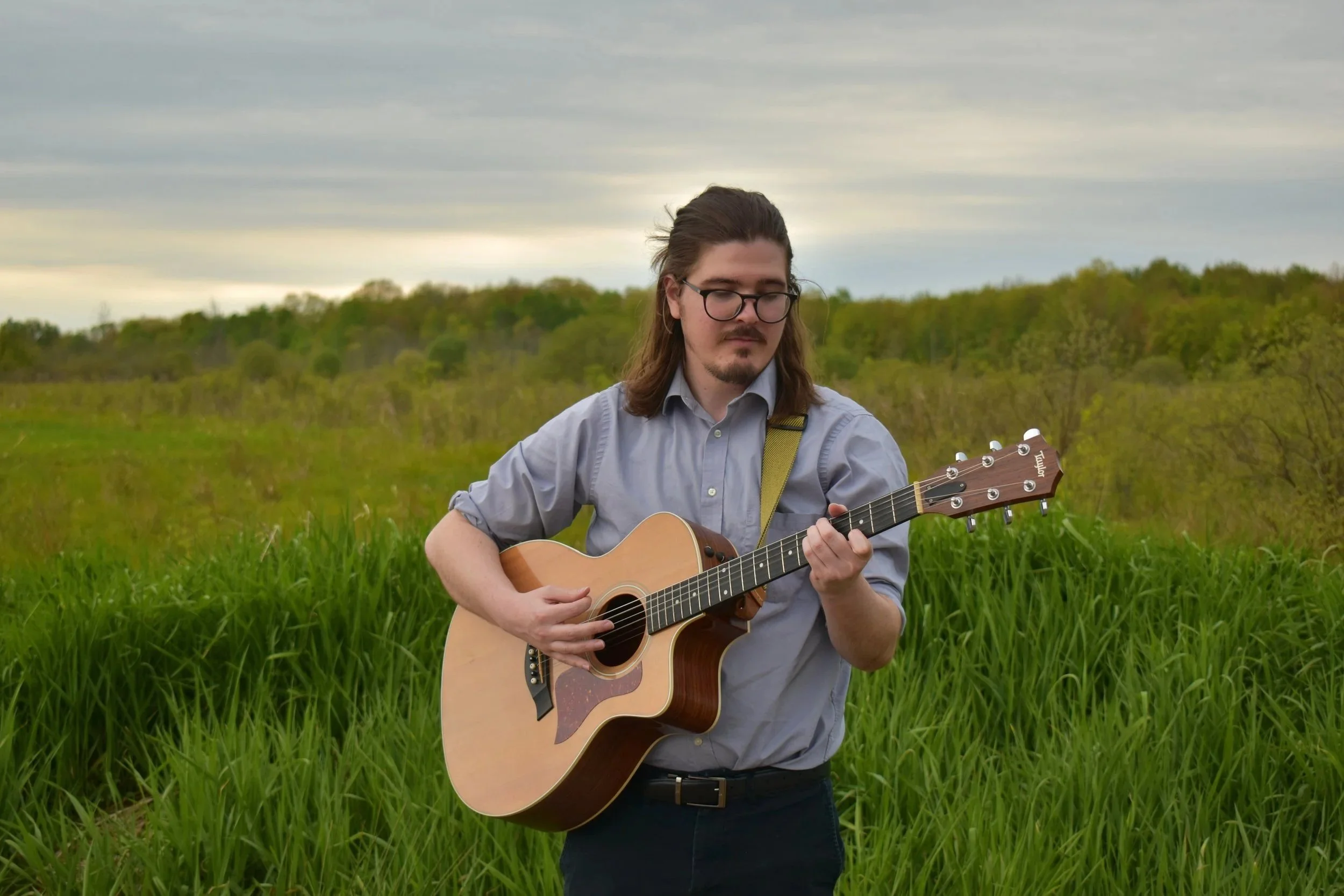 A man with long hair, glasses, and a mustache playing an acoustic guitar outdoors in a green field with trees and a cloudy sky in the background.