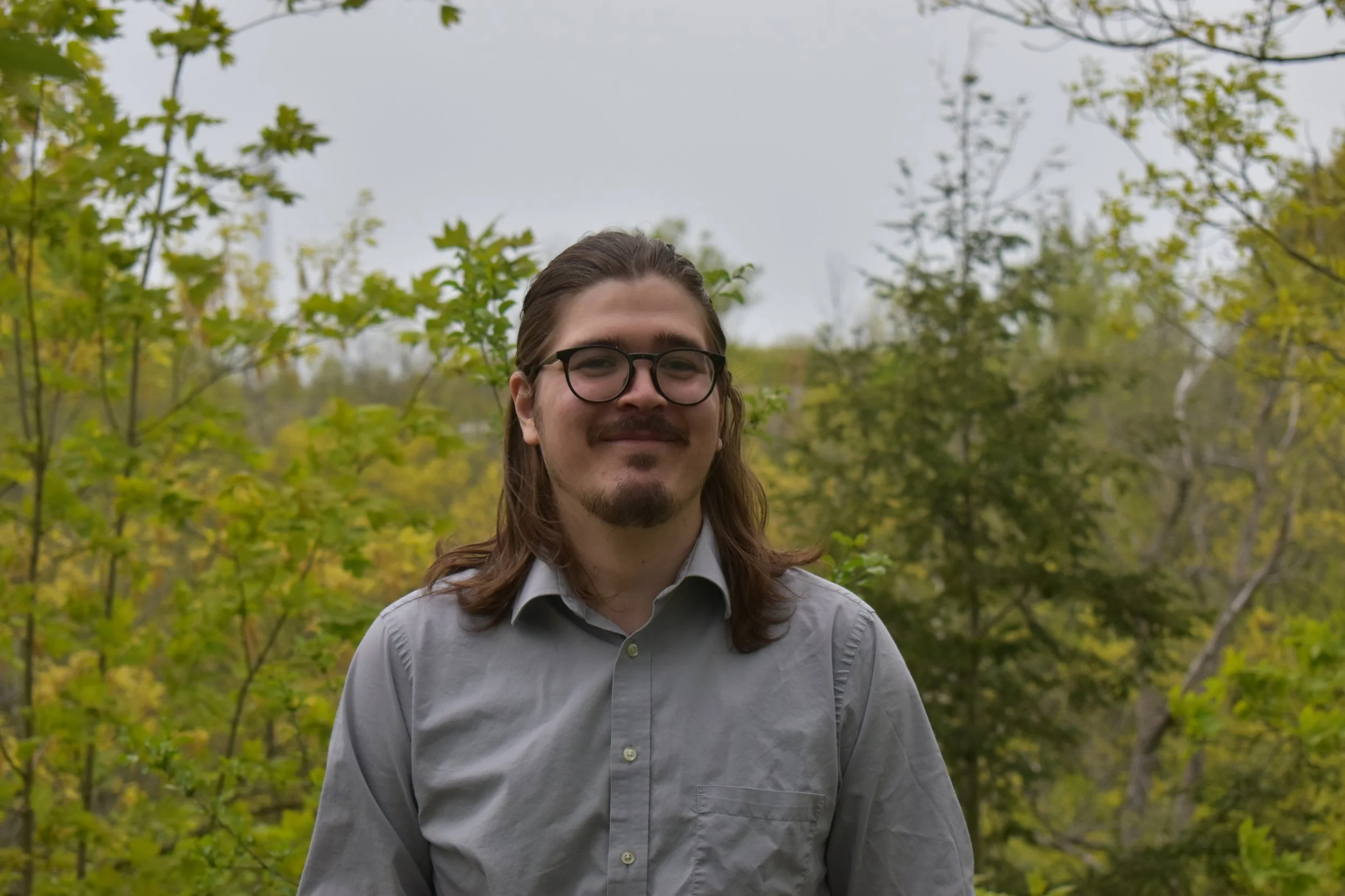 A man with long hair, glasses, and a mustache, wearing a light gray shirt, standing outdoors with trees and green foliage in the background.