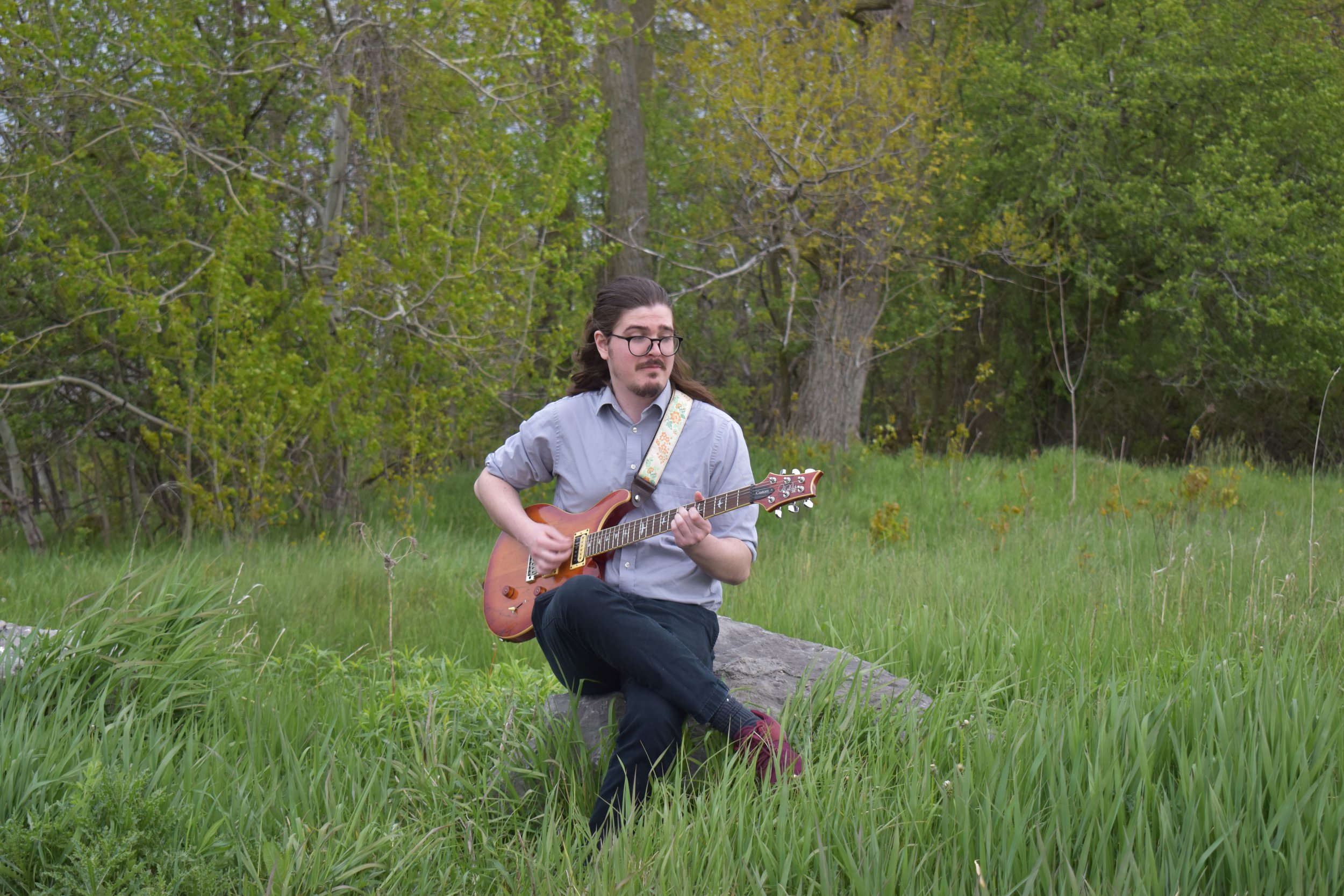 A man sitting on a rock in a grassy field playing an electric guitar, surrounded by green trees in the background.