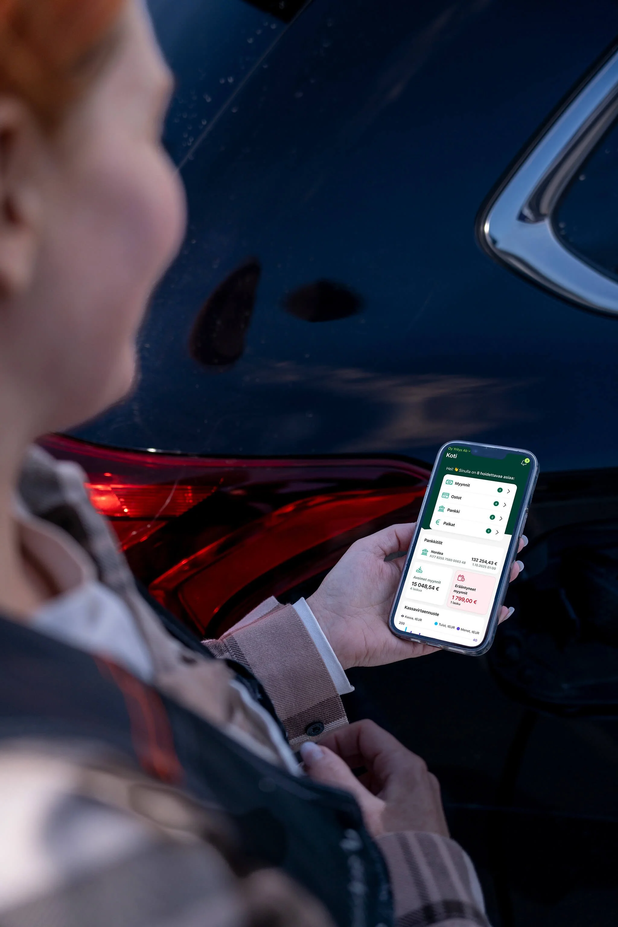 Henkilö, joka on istumassa autossa ja katsoo älypuhelintaan, jossa näkyy pankkisovellus. Vain osittainen näkymä auton takavalosta ja puhelimen näytöstä.