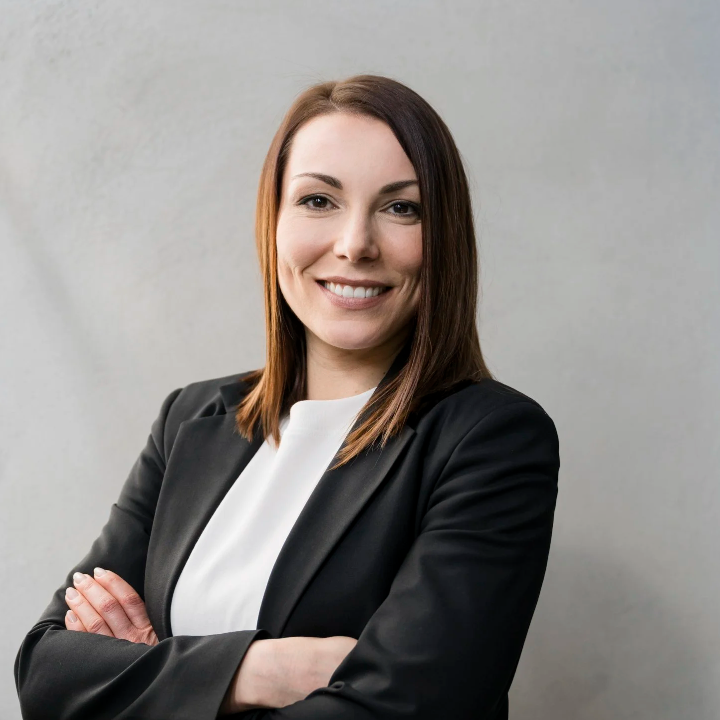 Eine lächelnde Frau mit braunen Haaren, die ein schwarzes Blazer und eine weiße Bluse trägt, steht vor einem grauen Hintergrund.