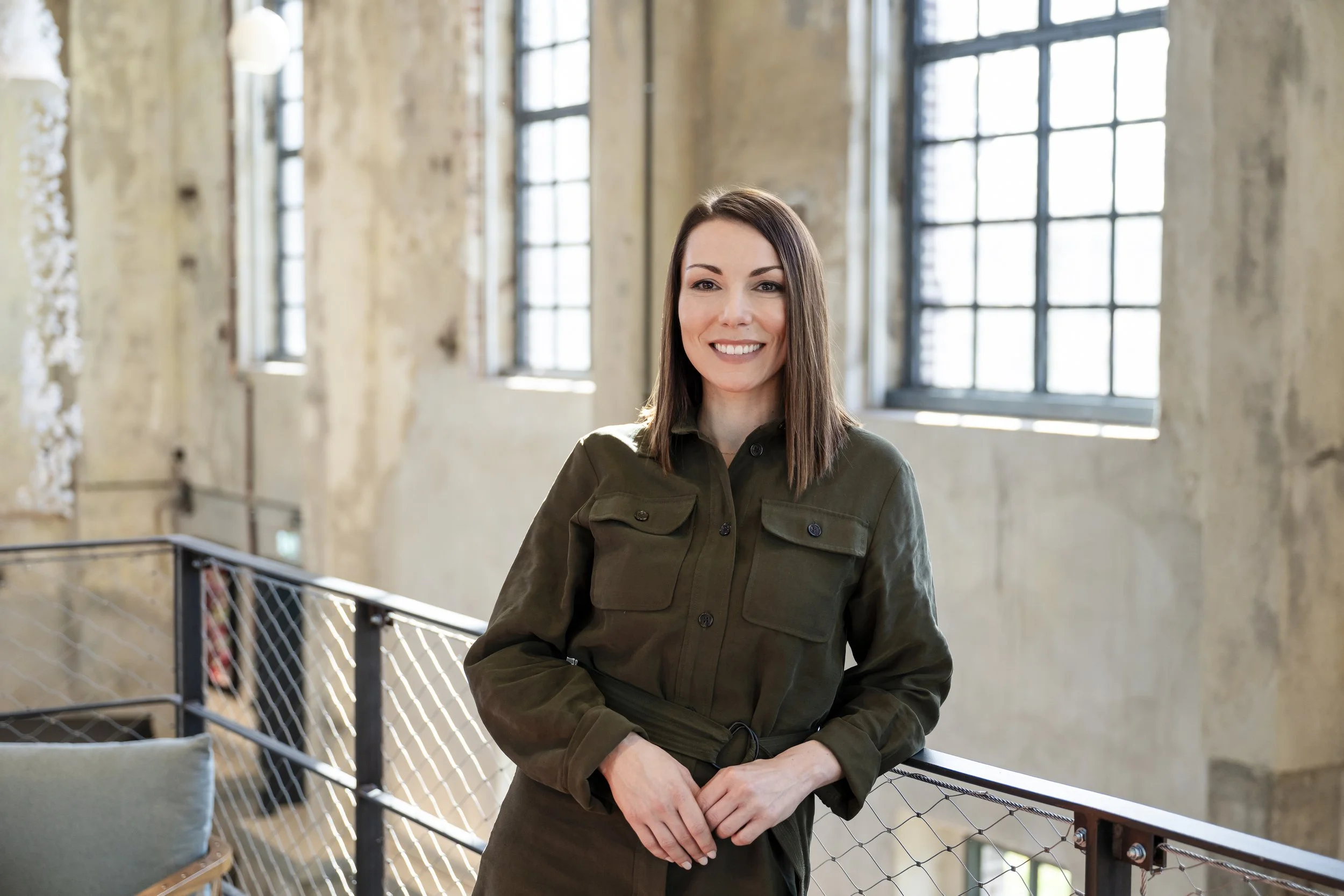 Junge Frau mit braunen Haaren, lächelnd, trägt grüne Jacke, steht in einer Industriehalle mit großen Fenstern und rohem Wänden.