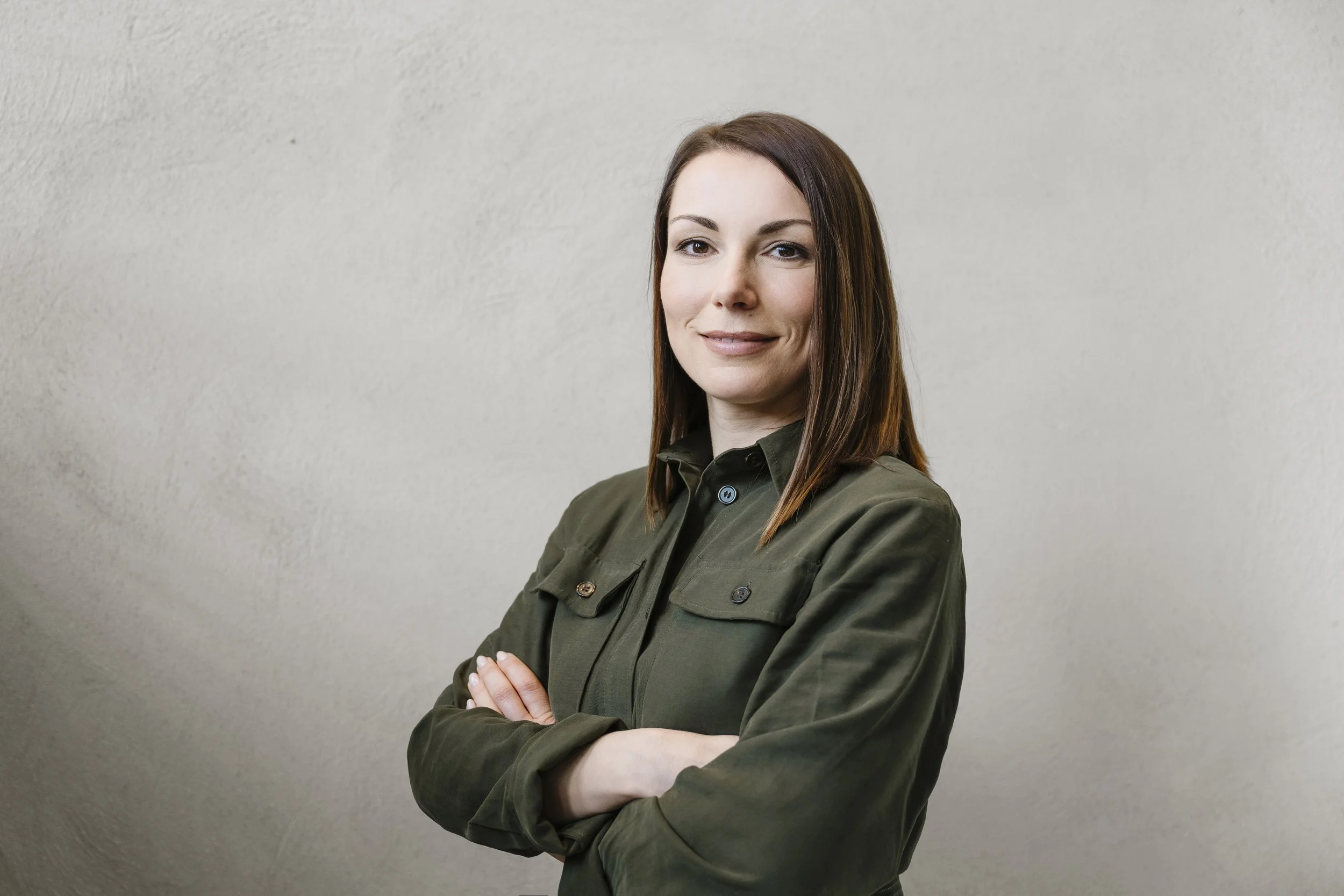 Frau mit braunen Haaren, grüner Jacke, posiert vor einer hellen Wand.