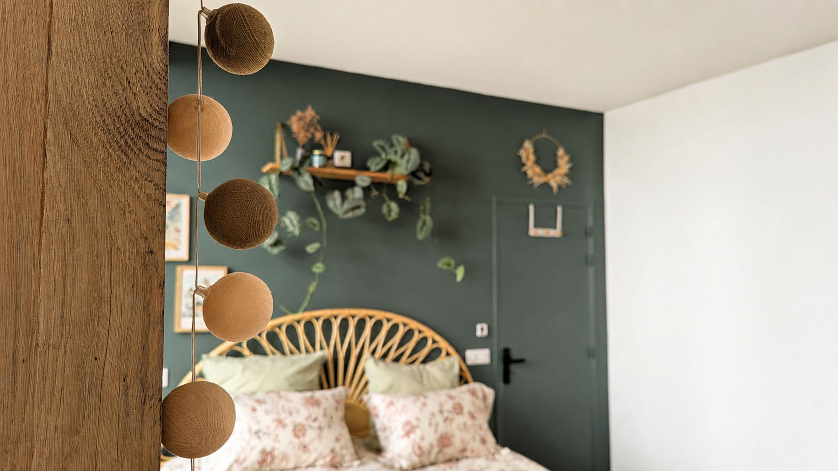 Décoration intérieure d'une chambre avec des coussins à motifs floraux, un lit en rotin, une porte verte foncée, un mur blanc, et une décoration en forme de couronne en bois. Hutch en bois avec des plantes suspendues et des cadres photo sur le mur