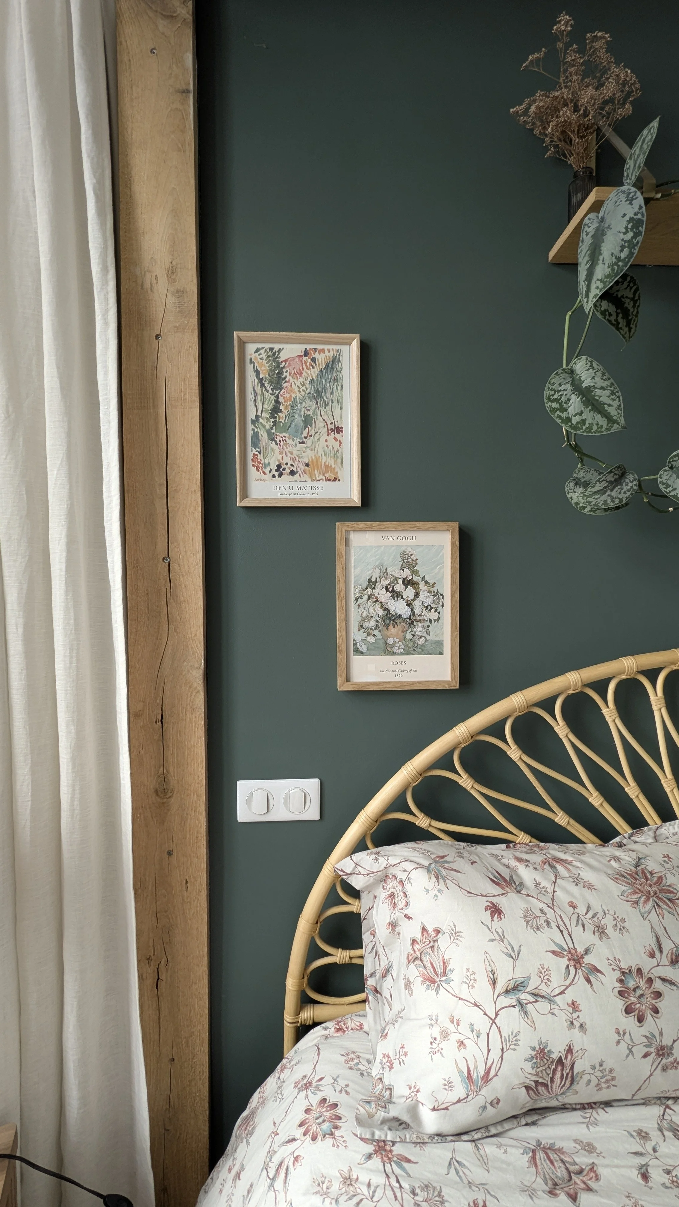 Coin chambre avec mur vert, deux cadres de posters d'art, une plante suspendue, un coin de lit avec une tête de lit en rotin et une parure de lit à motifs floraux, une prise électrique.