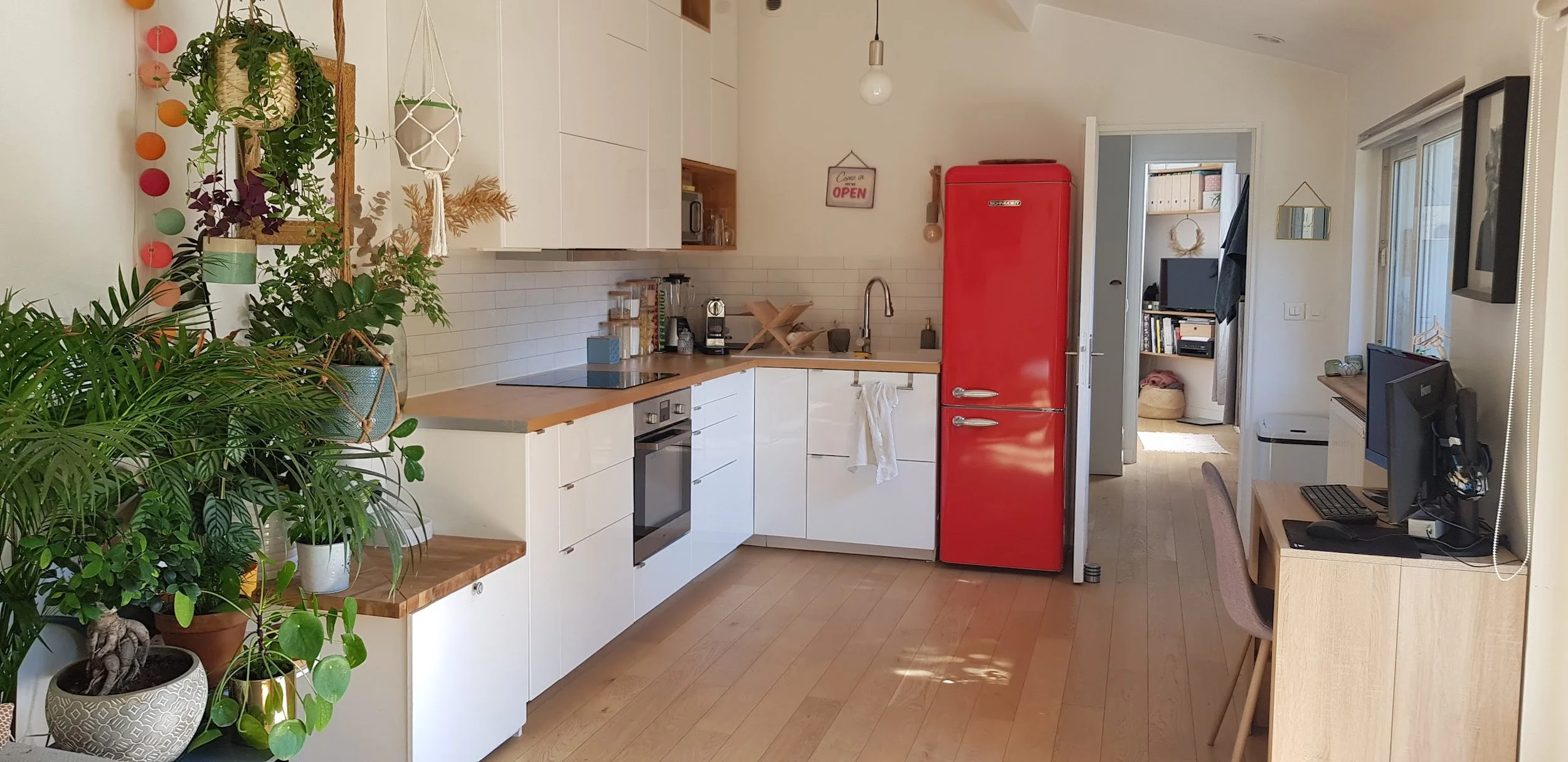 Cuisine moderne avec un réfrigérateur rouge, comptoir en bois, plants d'intérieur, et un espace de travail avec un ordinateur.