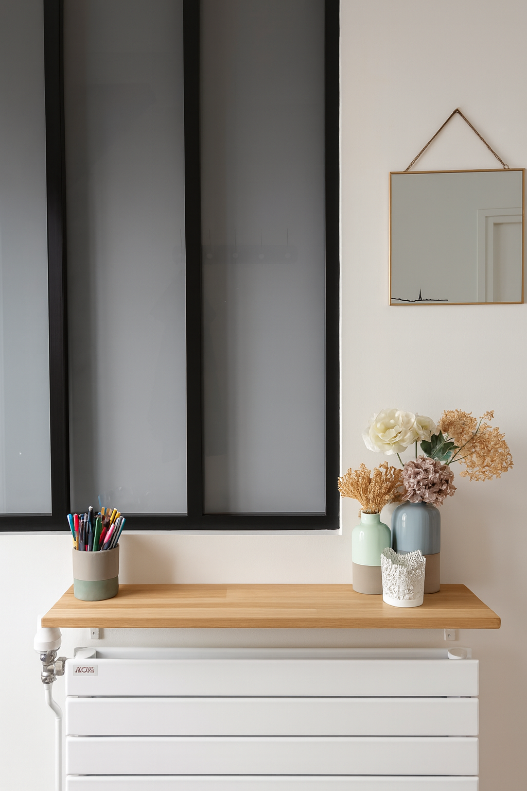 Une fenêtre en verre fumé, un miroir suspendu avec une chaîne dorée, et une table en bois avec des vases contenant des fleurs séchées et une boîte à crayons