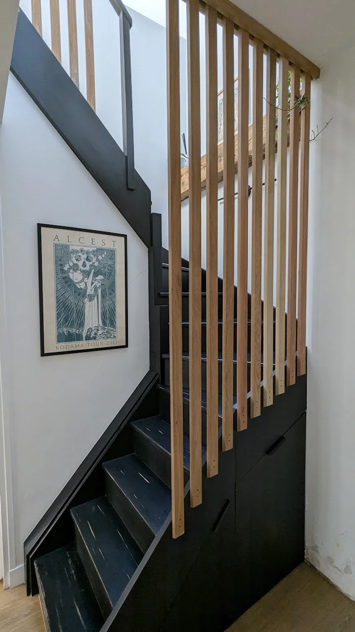 Escalier intérieur en bois et métal avec une paroi en lattes de bois verticales, une affiche encadrée sur le mur, et un sol en bois.