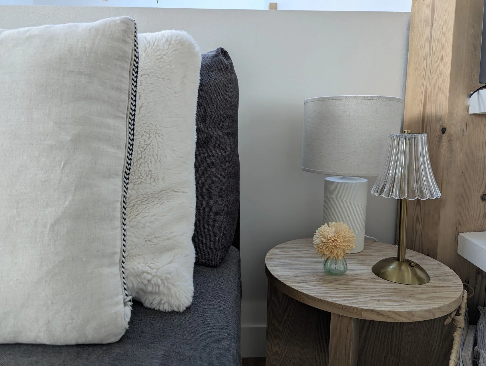 Un coin de canapé, des coussins, une table d'appoint avec une lampe, un vase avec une fleur, et une poutre en bois.