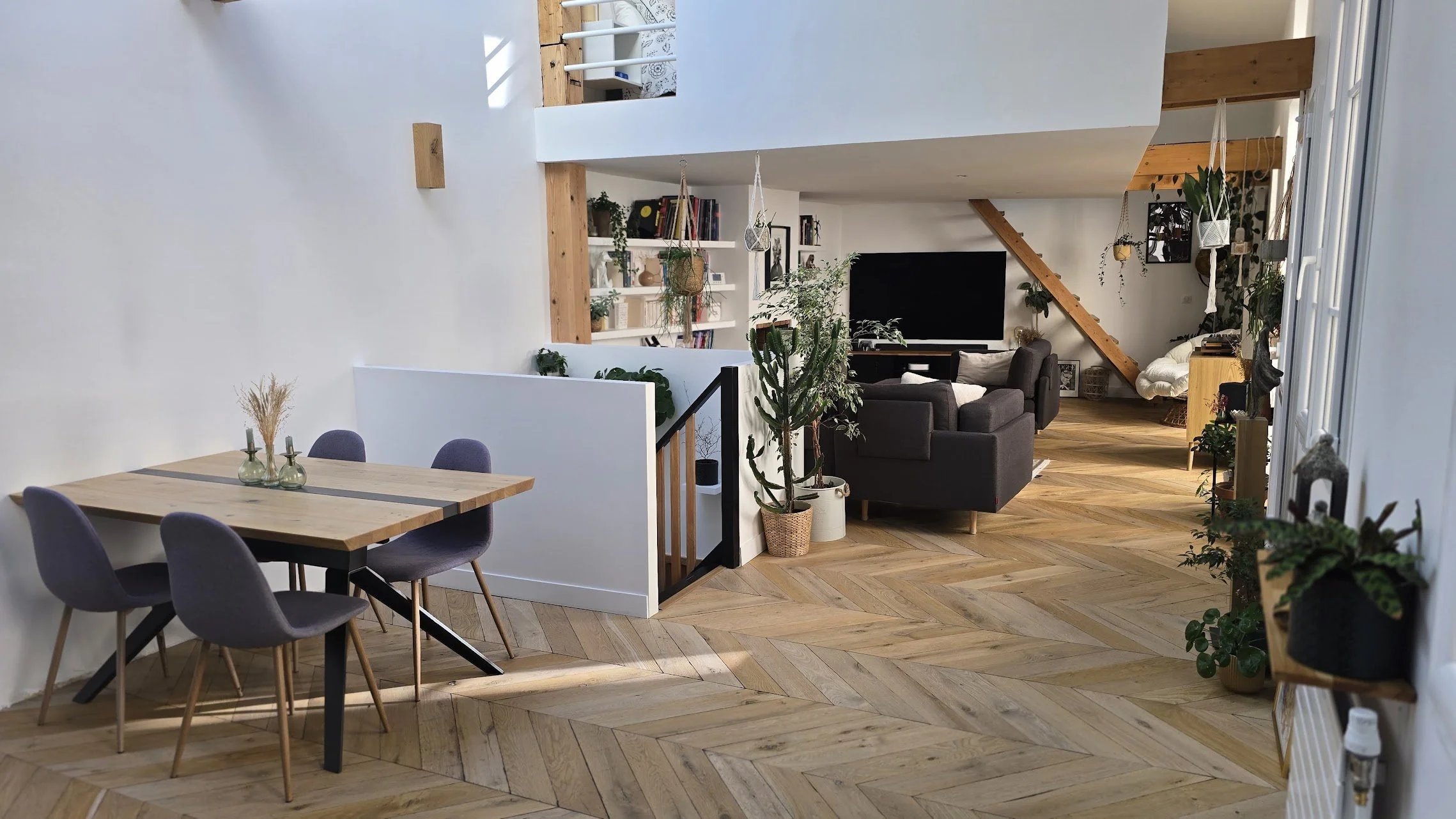 Intérieur d'un salon et salle à manger modernes avec mobilier en bois, plantes vertes, murs blancs et plancher en parquet. Espace lumineux et décoré avec des livres, des cadres et des éléments de décoration.
