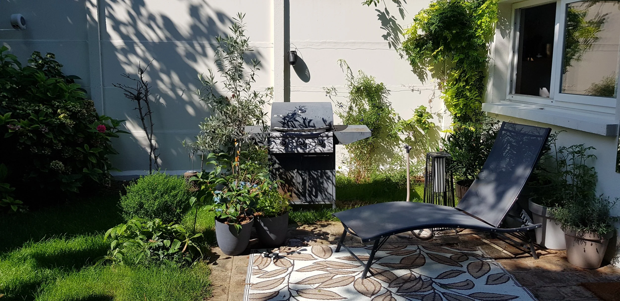 Petit jardin arrière avec un barbecue, un fauteuil pliant, plusieurs pots de plantes, un tapis de sol, et de la végétation verdoyante autour, avec une fenêtre de maison visible sur le côté droit.